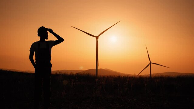 Silhouette Cinematic shot of young engineer putting security helmet on satisfied with his job on background of wind mills with at sunset.Concept: renewable energy, technology, electricity, service.