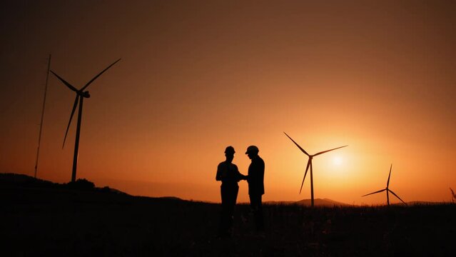 Silhouette Engineer and inspector working together on windmill farm. Engineer and indian inspector using digital tablet during meeting outdoors. Two partners standing on field with wind turbines.