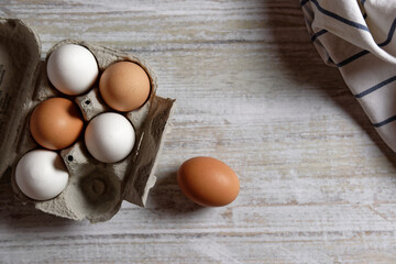 View from above of brown and white chicken eggs in an egg carton on gray wood. Next to it is a kitchen towel.