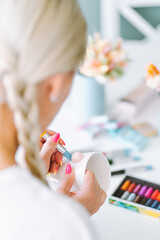 Vertical unrecognizable photo of woman artist hands with manicure jobbing and making decoration crafts flowers on mugs of polymer clay. Blurred color clay in box, cups with brushes, painters on table 