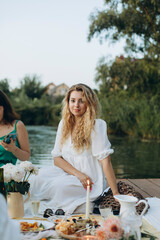 young beautiful blonde woman with wavy long hair sits on a wooden pier in a white dress. women's picnic party near the water. evening picnic dinner with snacks and burning candle.