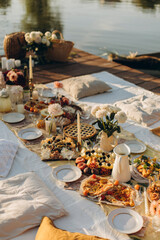 picnic on a wooden jetty near the water. a decorated picnic area awaiting guests. cheese and fruit plate on a picnic table. evening picnic party with golden sunlight.	