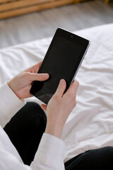Woman's hands hold a black tablet. Sits on a bed in an apartment.