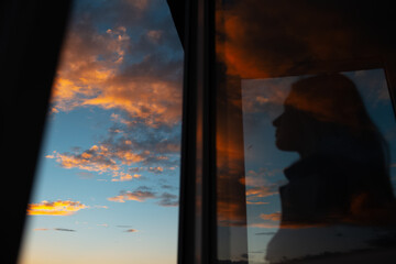 Blurred silhouette of girl through glass of window on background of sunset sky.