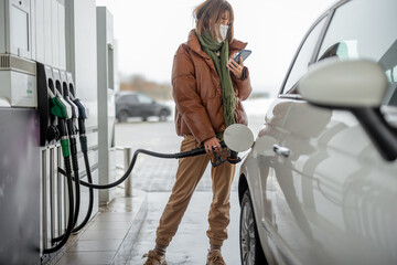 Woman in face mask refueling car with a gasoline, using smartphone to pay. Concept of mobile...
