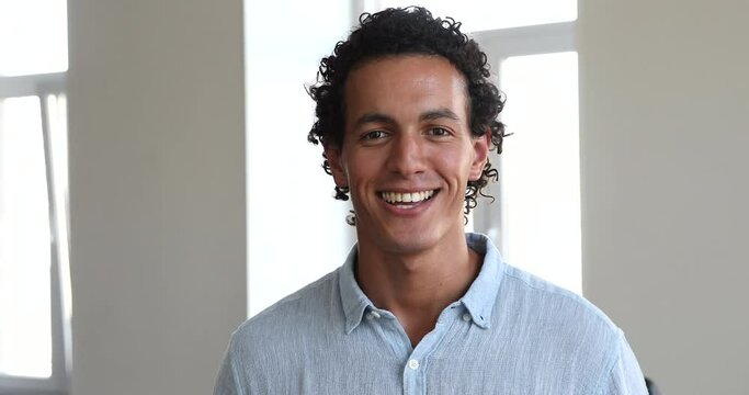 Handsome curly-haired Hispanic businessman in casual shirt standing alone indoor pose smile look at camera. Professional employee, advisory agency representative, counsellor head shot portrait concept