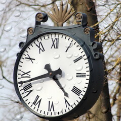A large round mechanical clock with Roman numerals in the park. A dial with arrows showing the time.