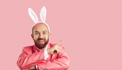 Look here, guys. Funny happy cheerful young man with ginger beard wearing pink suit and plush bunny ears pointing index finger to side at something on blank advertising copyspace Easter background