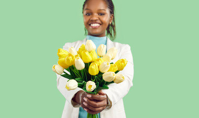 Happy Afro American girl holding a bunch of spring flowers and smiling. Closeup studio shot of a beautiful bouquet of fresh yellow and white tulips in the hands of a pretty young black woman - obrazy, fototapety, plakaty