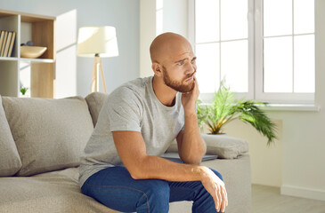 Upset young man holds his cheek and chin while suffering from toothache at home. Man with tired expression sits on sofa in living room and experiences severe toothache due to inflammation of nerve.