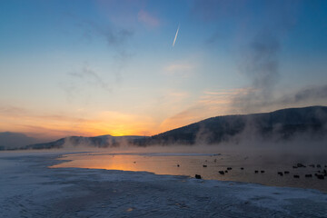 Winter blue landscape with the first orange rays of the sun over the unfrozen river. Beginning of dawn. Thick steam over water. Early morning on Yenisey river in Krasnoyarsk, Russia