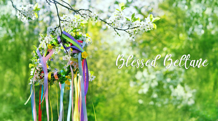 Blessed Beltane greeting card. Spring flower wreath with colorful ribbons in garden, green natural background. floral decor. Symbol of Beltane, Wiccan Celtic Holiday beginning of summer. witch ritual - obrazy, fototapety, plakaty