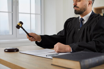 Serious young male judge in a robe uniform sitting at his table in the courtroom, banging the...