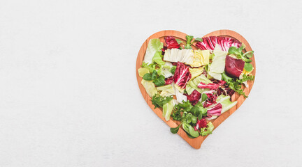 Fototapeta premium Sliced colored lettuce leaves in a wooden plate heart shape on a light cement background.