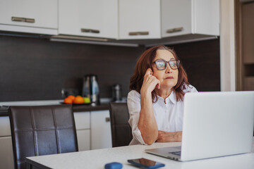 Beautiful pensive woman pensioner in glasses sitting at home at the table using a laptop