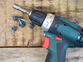 Close-up of a cordless screwdriver lying on a wooden surface with a nozzle for roofing screws. Self-tapping screws painted in gray are lying nearby. Top view, flat lay