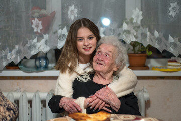 A girl hugs her old great-grandmother sitting on a chair.