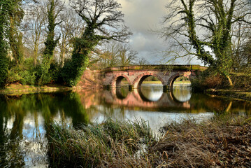 Fototapeta na wymiar Eine schöne alte Sandsteinbrücke spiegelt sich im Wasser eines See im Frühling in einer Parklandschaft