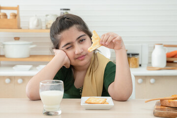 Little cute Indian kid girl holding and eating crackers for breakfast with a glass of milk, jam and bread in kitchen at home. A good diet is important to the growth of children concept