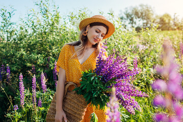 Woman put bouquet of lupin flowers in straw bag. Girl walking in summer meadow wearing yellow...