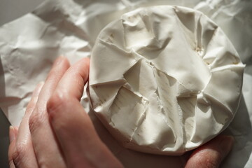 Fingers touch whole round camembert cheese on a sunny wrapping paper.