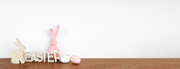 Easter decor on a wood shelf. Easter sign, eggs and modern glass bunnies against a white wall banner. Copy space.