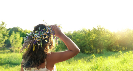 Girl in flower wreath on green sunny meadow, rear view. Floral crown, symbol of summer solstice. traditional Slavic ceremony on Midsummer, wiccan Litha sabbat. holiday Ivan Kupala. banner - obrazy, fototapety, plakaty