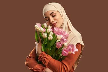 Portrait of a beautiful muslim woman wearing a beige hijab and holding a big bouquet of tulips on a brown background. The concept of female beauty, spring and femininity.