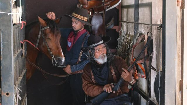 Two man with cowboy costume stay in horse stable that one check the bullet of long gun and other one take care horse in the back.