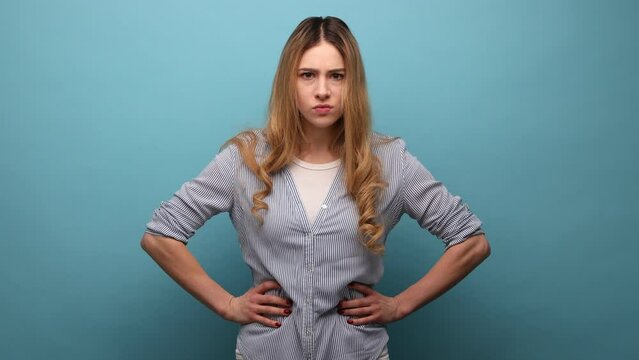 Portrait of young angry serious blond woman looking at camera with strict negative expression, aggression, holding hands on hips, wearing striped shirt. Indoor studio shot isolated on blue background.