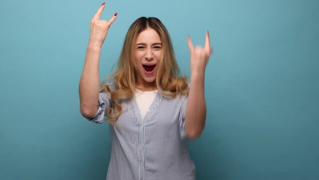 Portrait of overjoyed woman showing rock and roll hand sign, screaming and gesturing to heavy metal, rock music, wearing striped shirt. Indoor studio shot isolated on blue background.