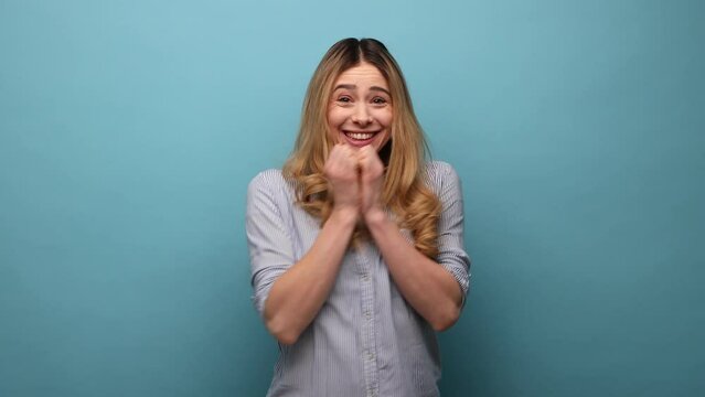 Portrait of shocked woman with wavy hair standing with raised arms and open mouth, hearing astonishing news, saying wow, wearing striped shirt. Indoor studio shot isolated on blue background.