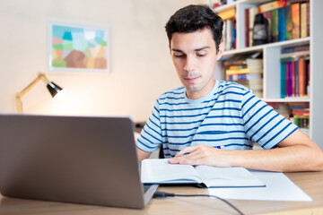 Student sitting at desk in his bedroom and using laptop to study, with book and paper by his side. Distance learning or e-learning through digital platforms.