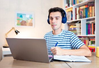 Student sitting at desk in his bedroom and using laptop to study, with book and paper by his side. Distance learning or e-learning through digital platforms.