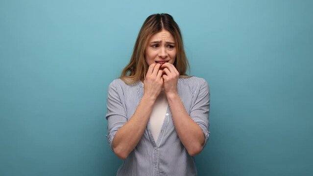 Troubles and worries. Portrait of nervous woman biting nails, terrified about problems, suffering phobia, anxiety disorder, wearing striped shirt. Indoor studio shot isolated on blue background.