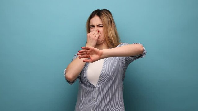 Bad smell. Portrait of young woman standing pinching her nose with fingers to hold breath, disgusted by stinky intolerable smell, wearing striped shirt. Indoor studio shot isolated on blue background.
