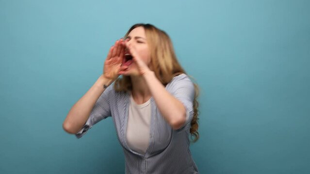 Portrait of angry yelling woman with wavy hair keeping hands near mouth, screaming loudly, expressing aggression, wearing striped shirt. Indoor studio shot isolated on blue background.
