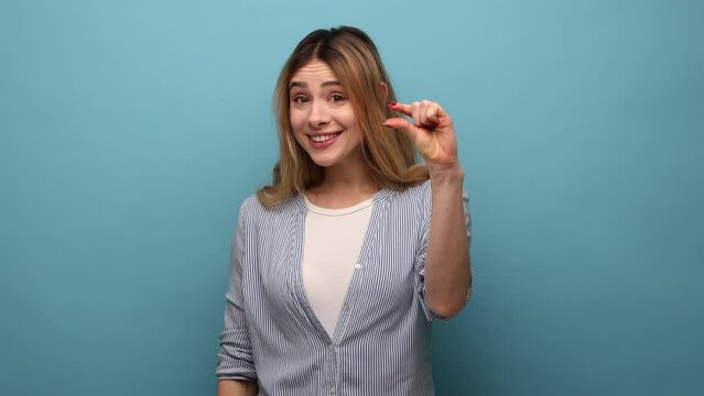 Upset frustrated woman with wavy hair showing small size with her fingers, sceptic about centimeter inch size, measuring scale, wearing striped shirt. Indoor studio shot isolated on blue background.