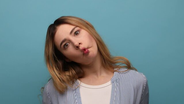 Funny surprised woman making fish face with pout lips and looking amazed, choking with idiotic comical silly expression, wearing striped shirt. Indoor studio shot isolated on blue background.