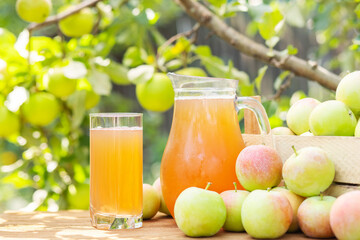 A box of apples and glass and pitcher of fresh apple juice on wooden table with garden background. Harvest of apples. Juice production