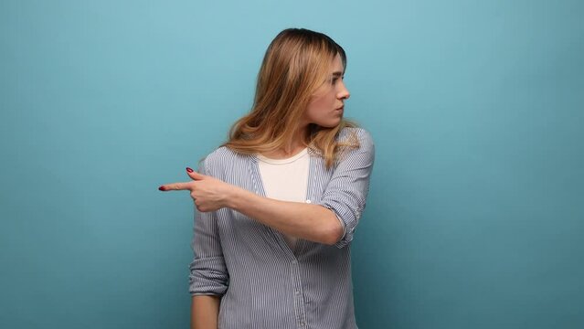 Upset vexed woman with wavy hair showing exit, demanding to leave her alone, turning away with resentful irritated expression, wearing striped shirt. Indoor studio shot isolated on blue background.