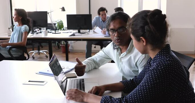 Multiethnic employees work in coworking office, Indian teammates colleagues sit at desk talking, share thoughts about online project development. Mentoring, collaboration, coworkers workflow concept