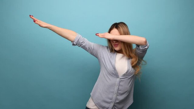 Portrait of attractive woman showing dab dance pose, famous internet meme of triumph, performing dabbing trends with hand gesture, wearing striped shirt. Indoor studio shot isolated on blue background