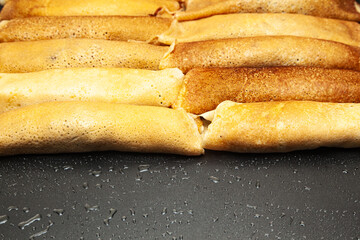 Fried and rolled pancakes are stacked side by side on a black, damp tray.