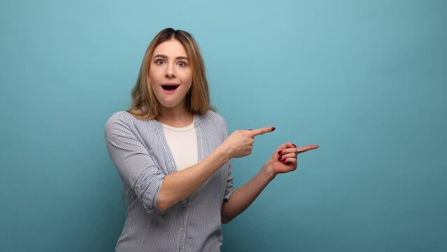 Crazy advertisement. Amazed shocked woman pointing to side copy space, gesturing attention to empty wall for commercial text, wearing striped shirt. Indoor studio shot isolated on blue background.