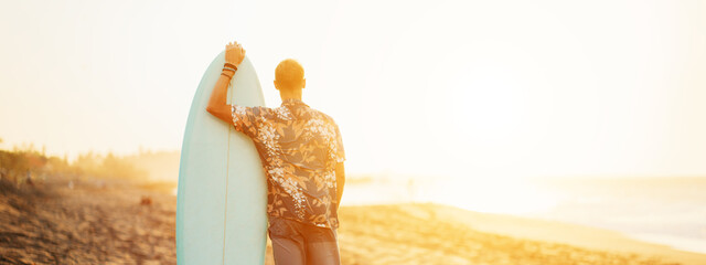 Portrait of happy surfer in hawaiian t-shirt with surf board on the beach, looking far away at the...