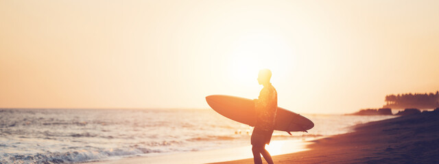 Surfer in hawaiian t-shirt with surf board on the beach at sunset, walking to the ocean, panoramic