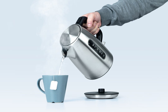 Male hand holding an electric kettle pouring hot water into a cup with a tea bag, isolated on a light blue background 