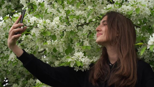 A teenager takes a selfie on a smartphone during the wind. A smiling young girl takes pictures of herself in the park against the background of a blooming white apple tree. High quality 4k footage