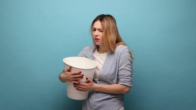 Sick unhealthy woman suffering abdominal cramps and nausea, vomiting, holding white bucket, symptoms of stomach flu or poisoning, wearing striped shirt. Indoor studio shot isolated on blue background.